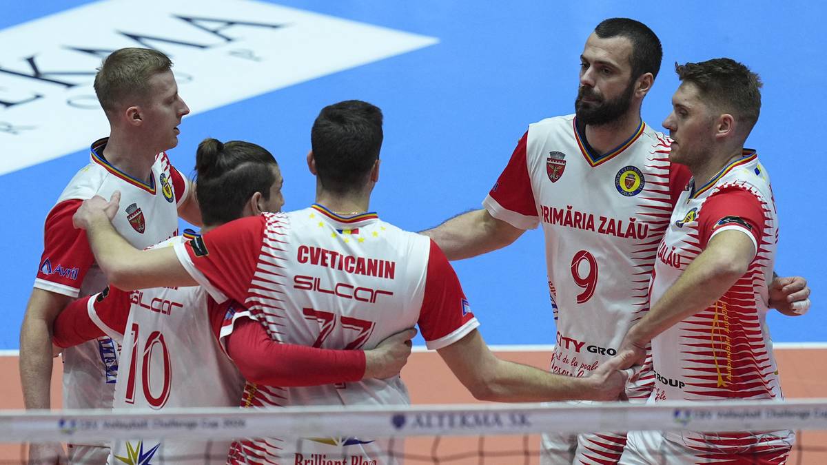 Drużyna siatkarzy w strojach z numerami na plecach, stojąca na boisku siatkarskim.