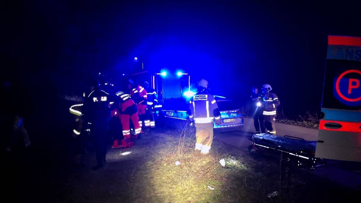 Grupa ratownik&oacute;w w mundurach, oświetlona niebieskimi światłami pojazd&oacute;w, pracuje na miejscu zdarzenia nocą. Widać nosze i fragment karetki.