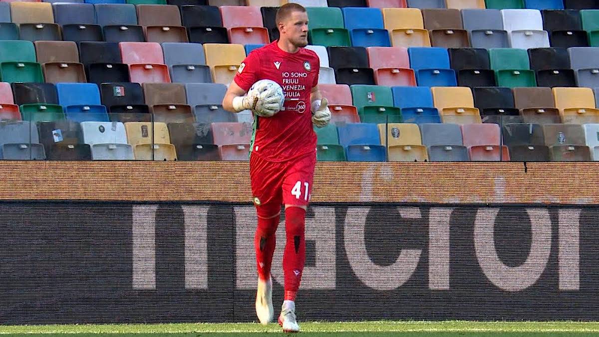 Bramkarz w czerwonym stroju sportowym z piłką w ręku, stojący na murawie stadionu z kolorowymi trybunami w tle.