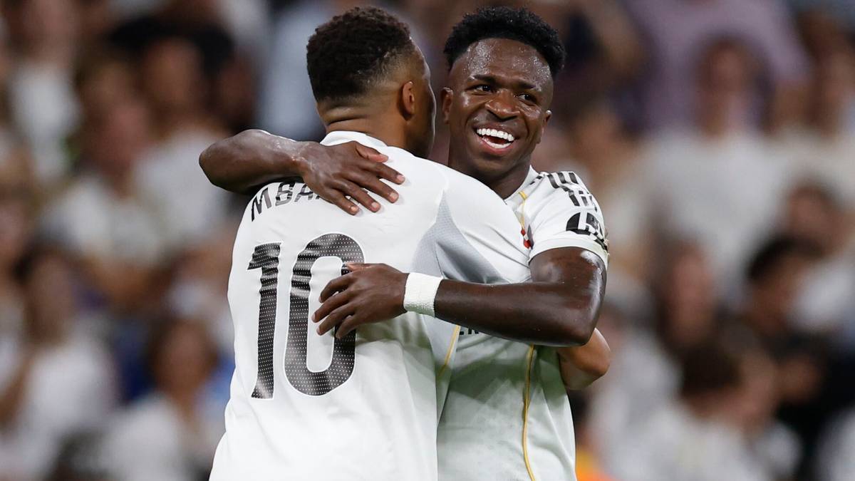 Dwóch piłkarzy Realu Madryt, Vinicius Jr i Kylian Mbappé, obejmują się po strzelonej bramce. Obaj ubrani są w białe stroje z numerami na plecach.