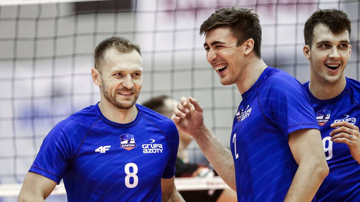 Dw&oacute;ch siatkarzy w niebieskich koszulkach rozmawia i śmieje się, trzeci siatkarz za nimi uśmiecha się do obiektywu.