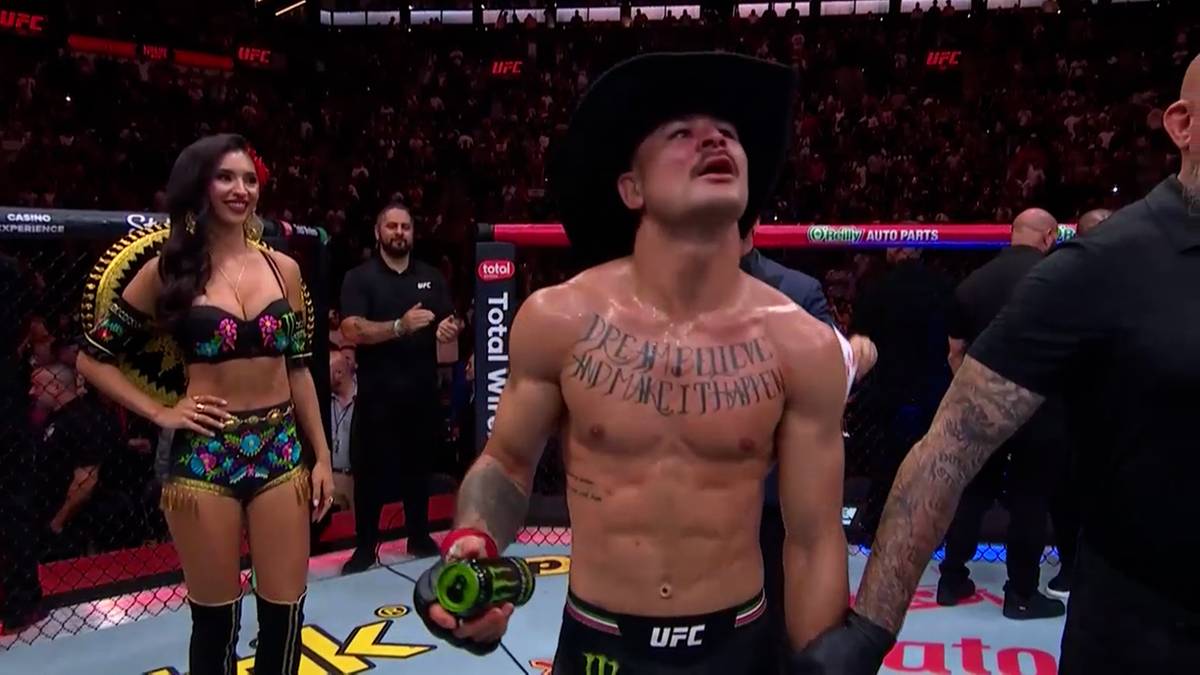 Mężczyzna w czarnym kapeluszu, z tatuażami na klatce piersiowej, stoi na ringu UFC, patrząc w górę. Obok niego po lewej stronie stoi kobieta w kolorowym stroju.