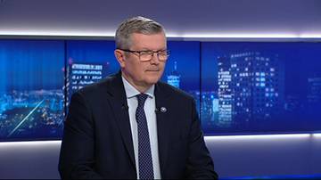 Mężczyzna w garniturze i okularach siedzi w studiu telewizyjnym na tle nocnego miasta.