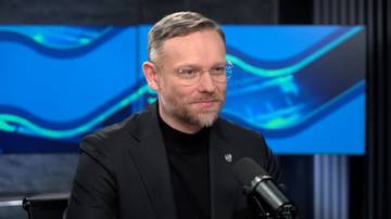 Mężczyzna w okularach, ubrany w czarny golf i marynarkę, siedzi i patrzy w prawo. Przed nim znajduje się mikrofon.