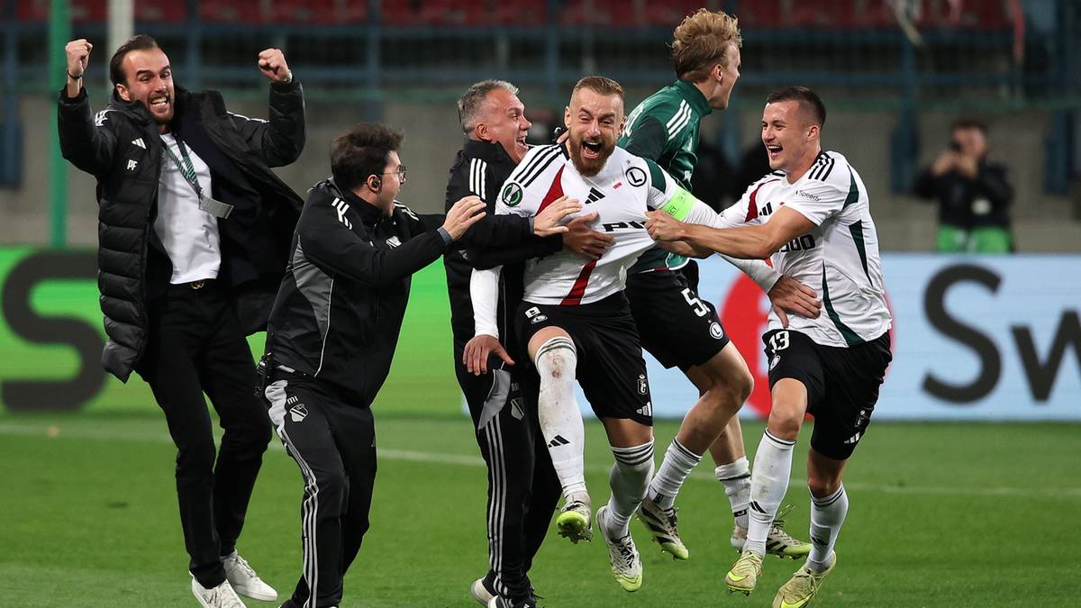 Piłkarze Legii Warszawa świętujący zwycięstwo na boisku, jeden z nich uniesiony na rękach przez kolegów.