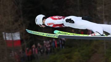 Skoczek narciarski w białym kombinezonie wykonuje skok, patrząc w d&oacute;ł.