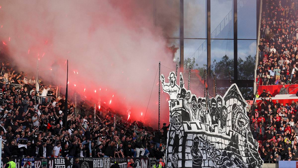Tłum kibiców na stadionie, z których część używa rac, wytwarzając czerwony dym. Na pierwszym planie widoczny jest duży czarno-biały transparent przedstawiający zamek z postaciami. W tle znajdują się trybuny wypełnione ludźmi i nowoczesna architektura stadionu.