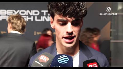 Portret młodego mężczyzny z ciemnymi włosami, udzielającego wywiadu na tle napisu "BEYOND THE FINISH LINE".