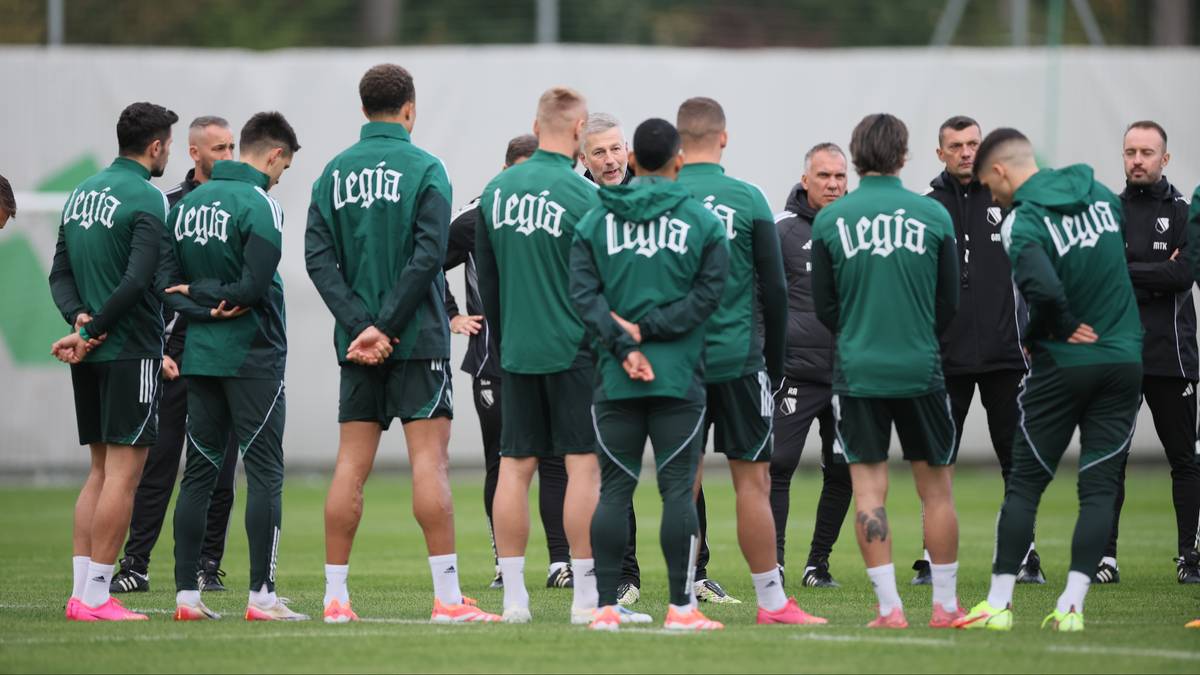 Grupa piłkarzy Legii Warszawa w zielonych strojach treningowych stoi w okręgu i rozmawia.