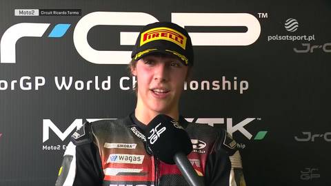 Młody zawodnik motocyklowy w czapce z daszkiem i stroju wyścigowym, z mikrofonem przed twarzą, podczas wywiadu po zawodach.