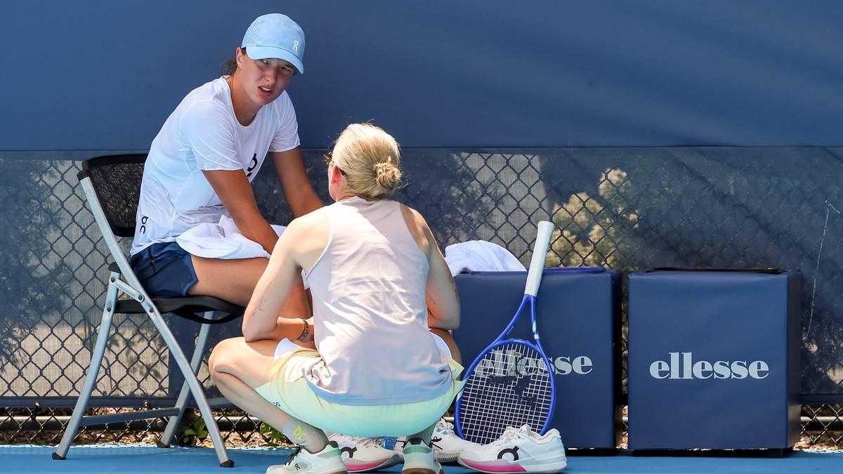 Mężczyzna w białej koszulce i czapce siedzi na krześle, rozmawiając z kobietą kucającą przed nim. Obok nich leży rakieta tenisowa i dwie niebieskie torby z logo "ellesse".