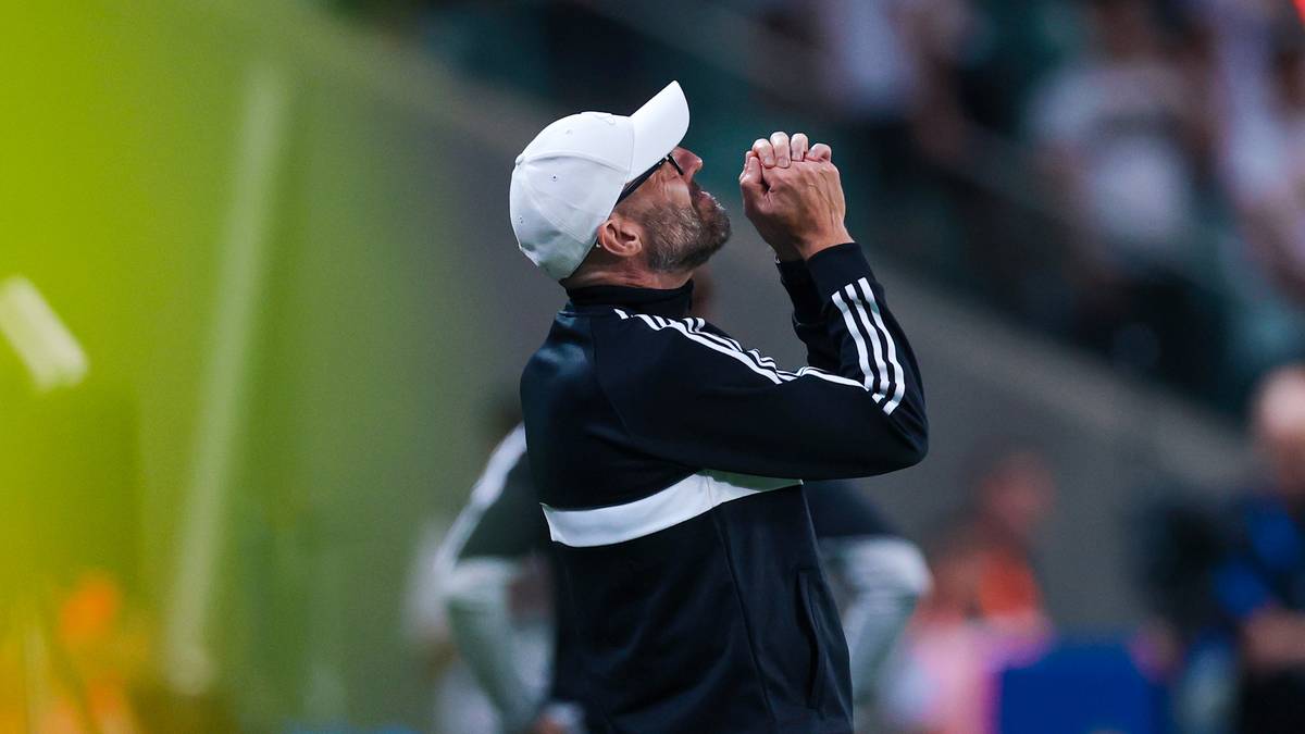 Mężczyzna w białej czapce z daszkiem i czarnej kurtce sportowej z białymi pasami na rękawach, ze złożonymi dłońmi przy twarzy, patrzący w g&oacute;rę z zamyśloną miną.