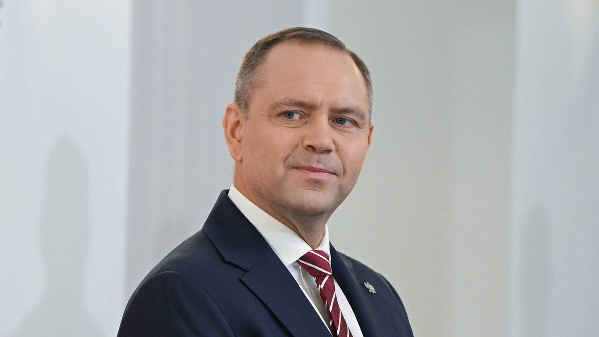 Grupa mężczyzn w garniturach stoi w rzędzie, z flagami Polski w tle.