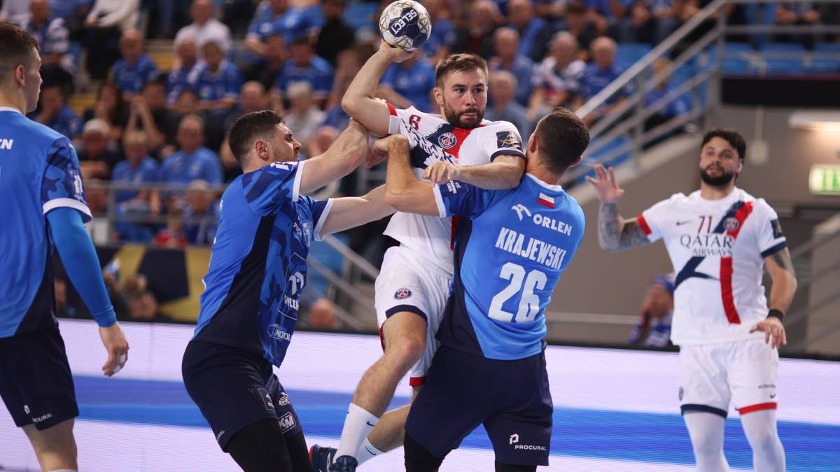 Piłkarz ręczny w białej koszulce z numerem 11 próbuje rzucić piłkę, podczas gdy trzech zawodników w niebieskich koszulkach z numerami 26 i 15 próbuje go powstrzymać. Na drugim planie widoczni są kibice na trybunach.