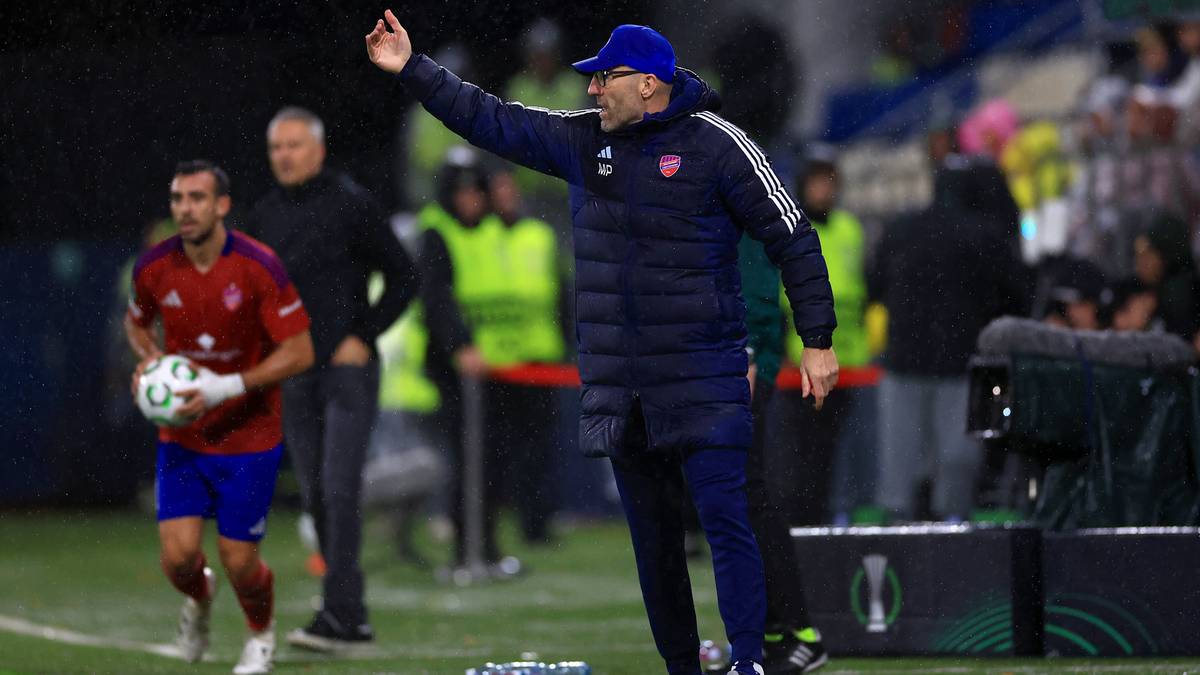 Trener piłkarski w czapce i kurtce gestykuluje na boisku, obok biegnie zawodnik z piłką.