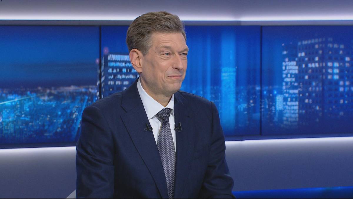 Mężczyzna w granatowym garniturze i białej koszuli z krawatem siedzi w studiu telewizyjnym.