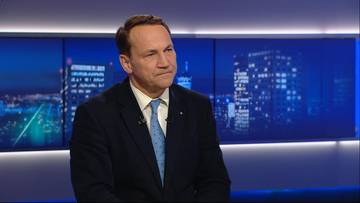 Radosław Sikorski siedzi w studiu telewizyjnym z widokiem na nocne miasto w tle.
