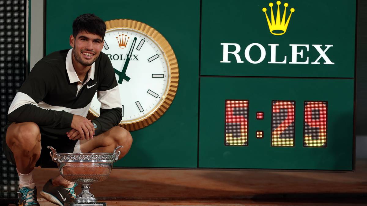 Hiszpański tenisista Carlos Alcaraz klęczy przed pucharem w towarzystwie zegara z logo Rolex i wyświetlacza pokazującego czas 5:29.
