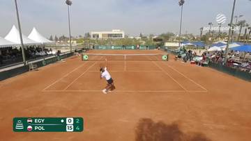 Gracz tenisa w białej koszulce i czarnych spodenkach wykonuje serwis na ceglanym korcie. Na ekranie widoczny jest wynik meczu: Egipt 0-2, Polska 15-4.