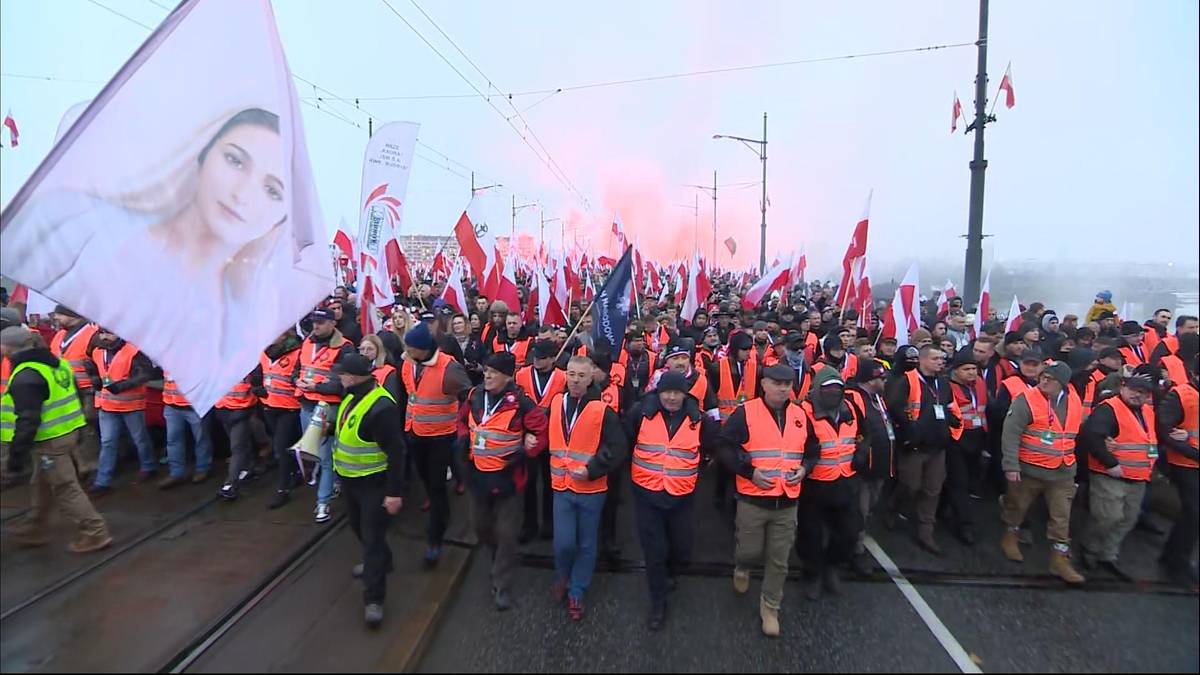 Grupa osób w kamizelkach odblaskowych z transparentami i flagami podczas marszu.