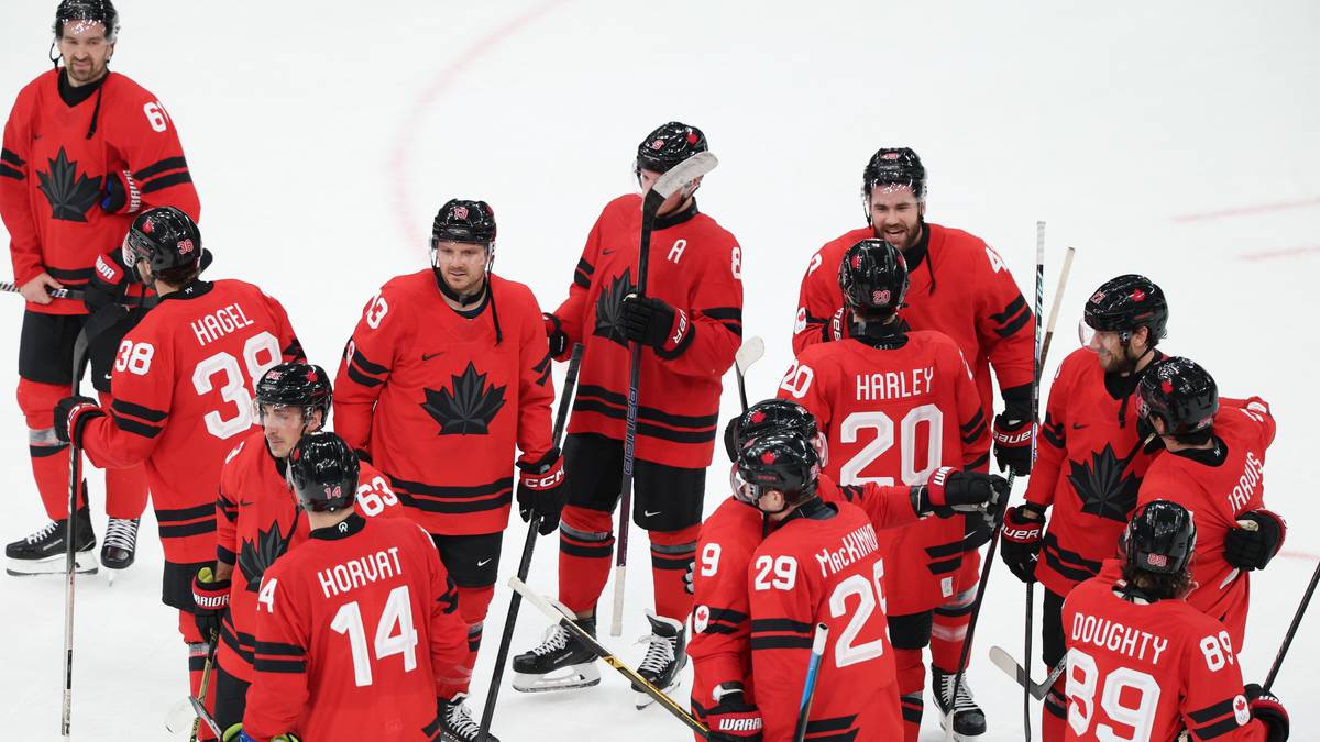 Grupa kanadyjskich hokeist&oacute;w w czerwonych strojach z flagą narodową na piersiach i numerami na plecach stoi na lodowisku, trzymając kije.