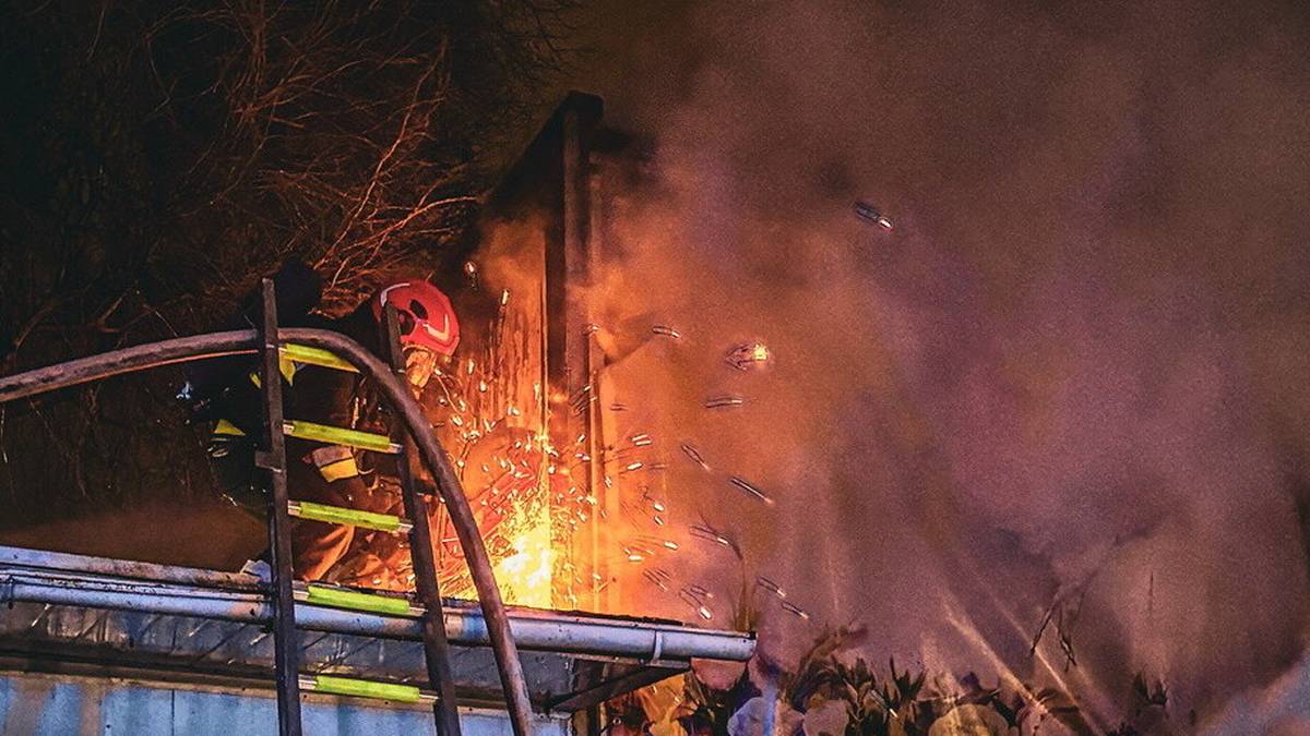 Strażak w hełmie i ubraniu ochronnym kieruje strumień wody na płonący dach budynku, otoczony dymem.