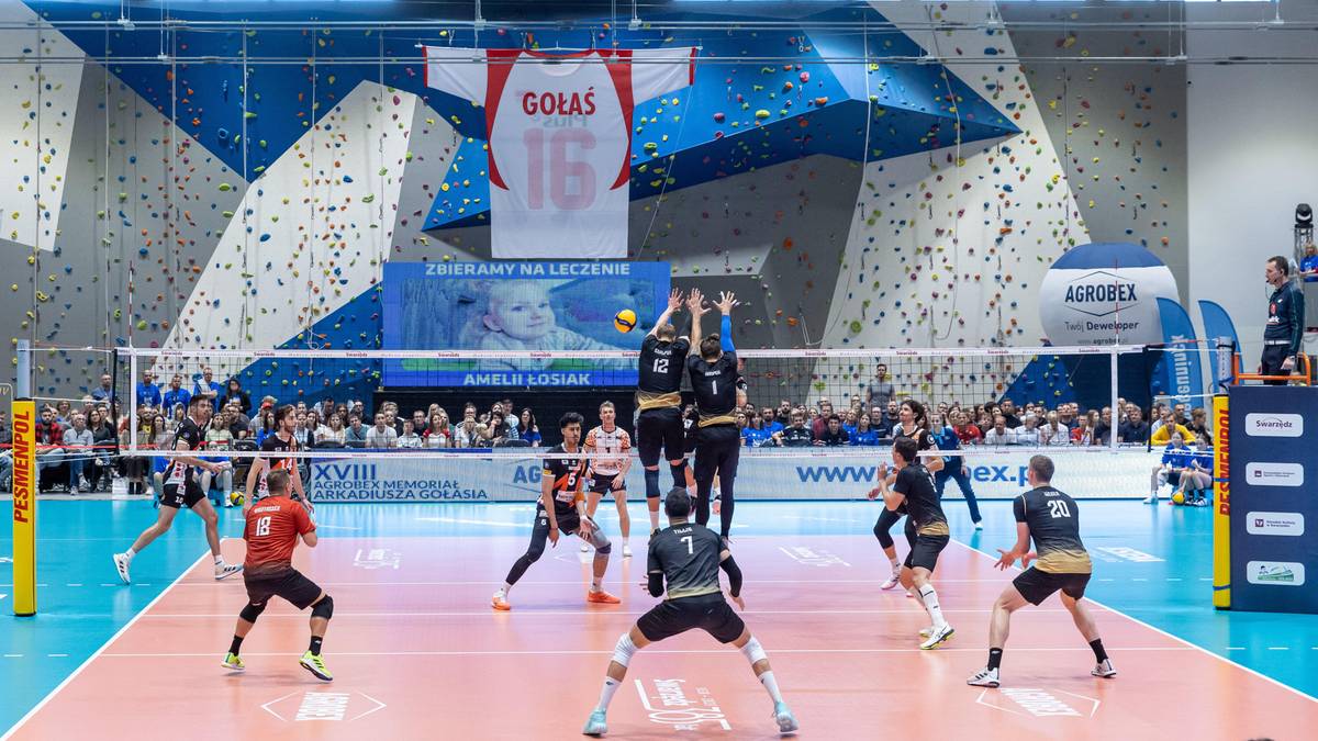 Mecz siatkówki w hali sportowej z zawodnikami po obu stronach siatki. Na trybunach siedzą widzowie. Nad boiskiem wisi baner z napisem "GOŁAŚ" i numerem "16".