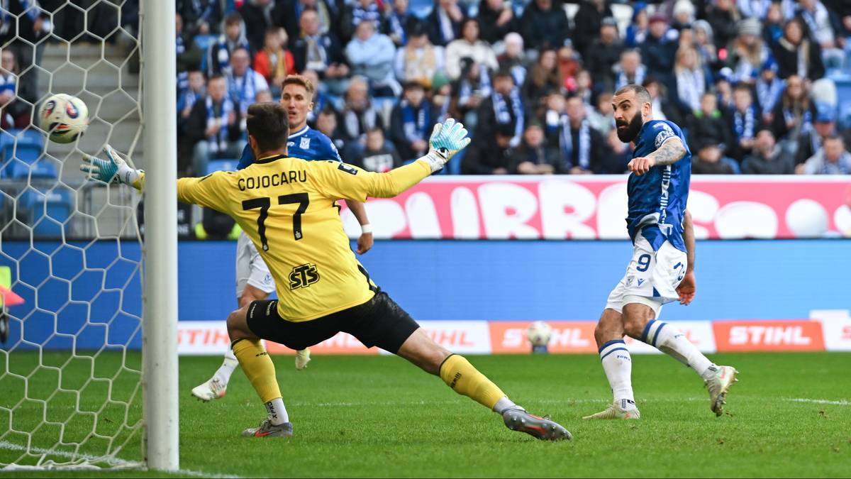 Piłkarz w niebieskim stroju strzela bramkę, bramkarz w żółtym stroju próbuje interweniować.