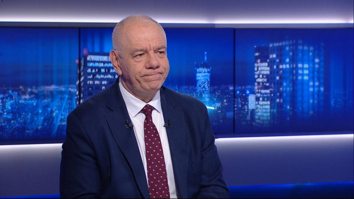 Jacek Sasin w ciemnym garniturze i białej koszuli z krawatem, siedzi przed ekranem z widokiem nocnego miasta.