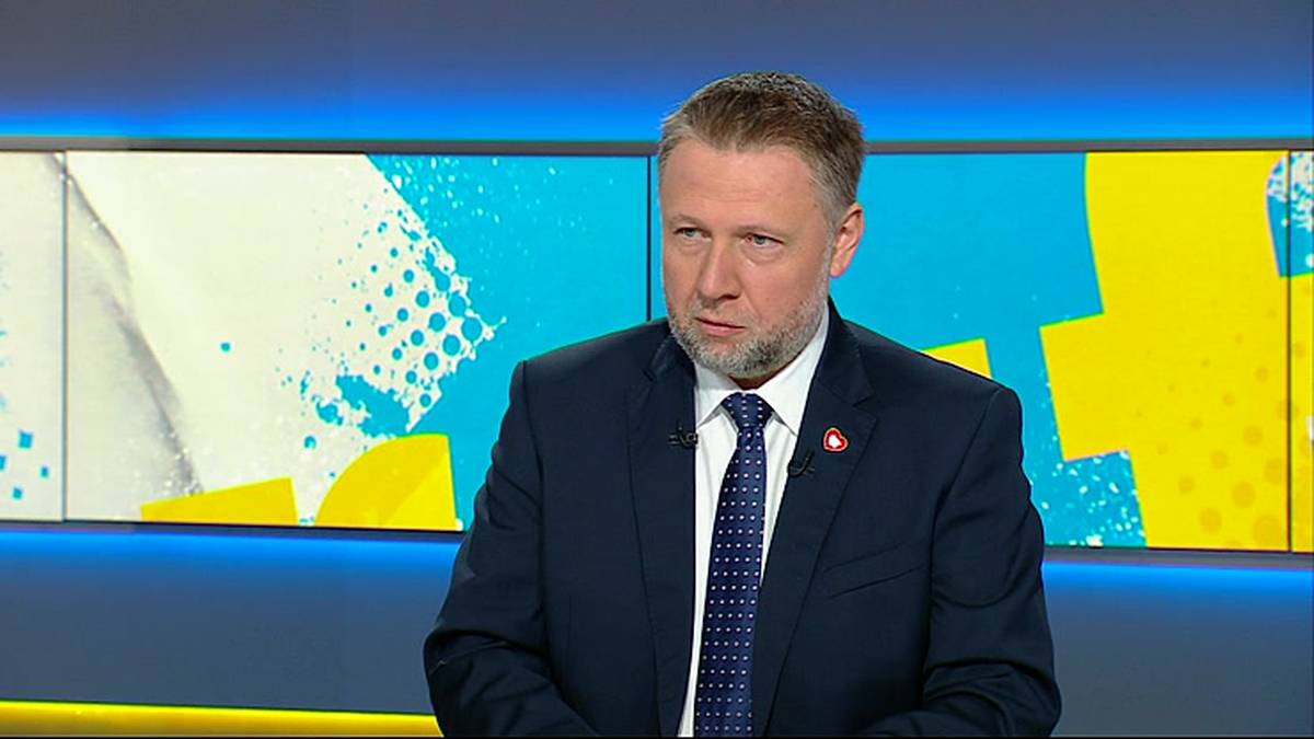 Mężczyzna w średnim wieku w granatowym garniturze i krawacie, siedzi przed ekranem z kolorowymi abstrakcyjnymi wzorami.