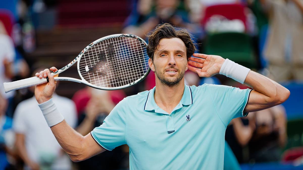 Tenisista w niebieskiej koszulce z krótkim rękawem, opaską na nadgarstku i rakietą tenisową w dłoni, z uśmiechem na twarzy, na tle publiczności.