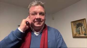 Mężczyzna w średnim wieku, ubrany w granatowy sweter i czerwony szalik, opiera prawą dłoń o ucho i patrzy w stronę obiektywu. W tle widoczny obraz w ramie.