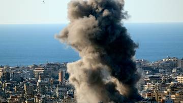 Ogromny słup dymu unoszący się nad zabudowanym miastem, widoczne morze w tle.