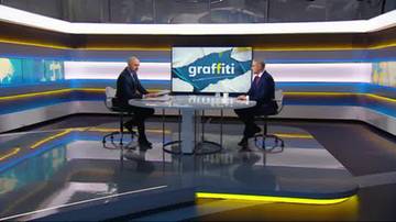 Dw&oacute;ch mężczyzn w garniturach siedzi przy okrągłym stole w nowoczesnym studiu telewizyjnym. Na ekranie za nimi widnieje napis "graffiti". Jeden z mężczyzn wskazuje na ekran.