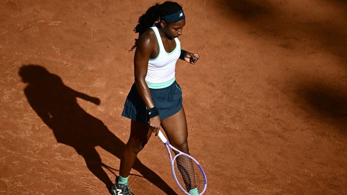 Tenisistka Coco Gauff w białej koszulce i granatowej sp&oacute;dniczce idzie po korcie ziemnym z rakietą w ręku, patrząc w d&oacute;ł.