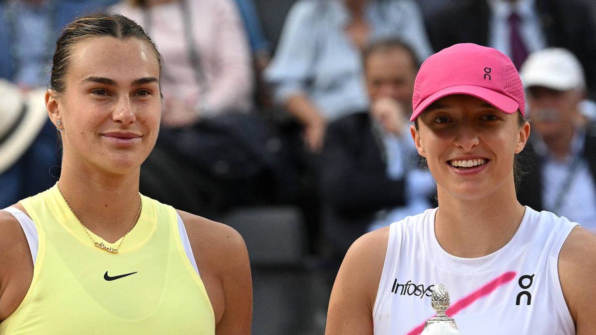 Dwie tenisistki, Iga Świątek w białej koszulce i różowej czapce z daszkiem, oraz Aryna Sabalenka w żółtej koszulce, patrzą w stronę obiektywu z lekkim uśmiechem. W tle widać niewyraźne postacie widzów.