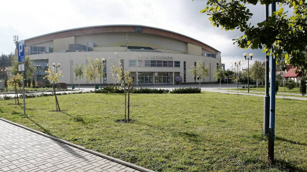 Zdjęcie przedstawiające nowoczesną halę widowiskowo-sportową z przeszkloną fasadą, otoczoną zielonym trawnikiem i drzewami.