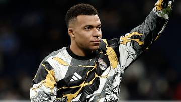 Piłkarz Kylian Mbappe w bluzie treningowej z charakterystycznym wzorem, macha do publiczności na stadionie.