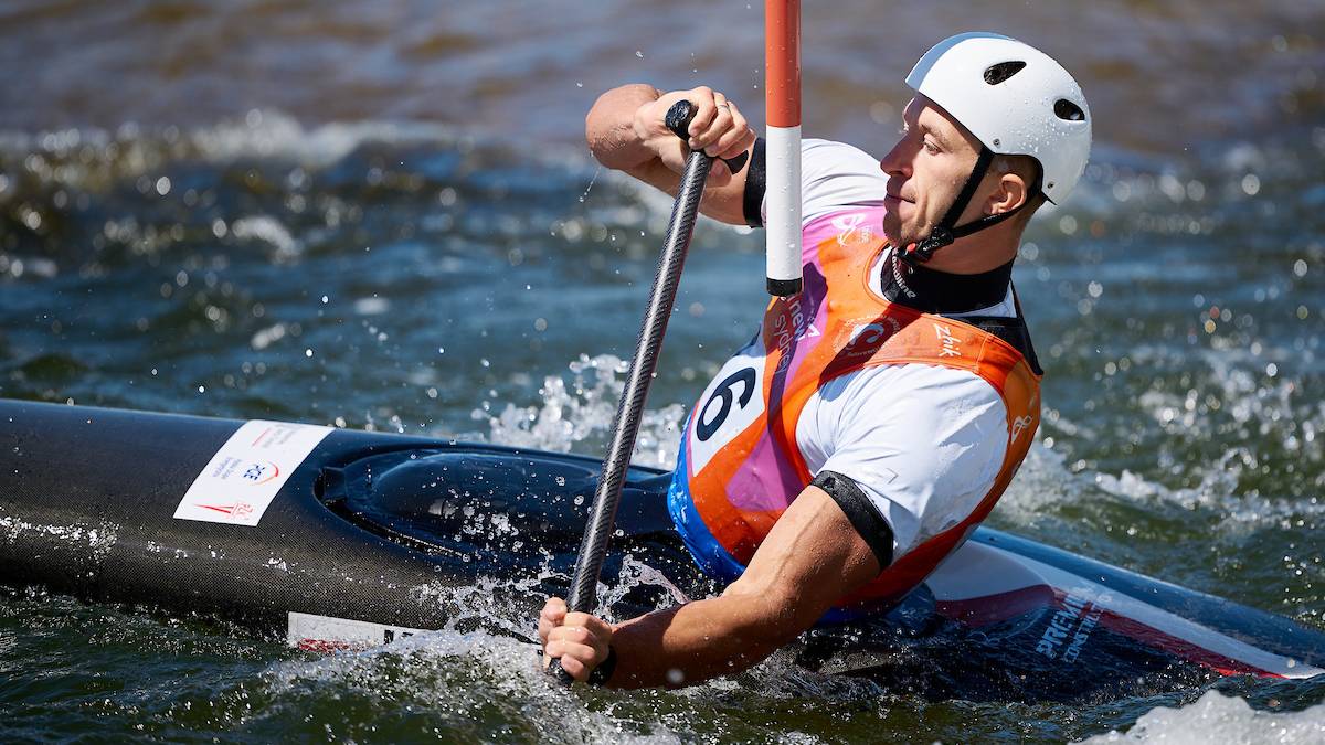 Mężczyzna w białym kasku i kamizelce ratunkowej wiosłuje w kajaku po wzburzonej wodzie.