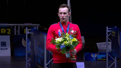 Łukasz Gutkowski na podium, trzymający bukiet kwiat&oacute;w i puchar, ubrany w czerwoną bluzę z medalem.