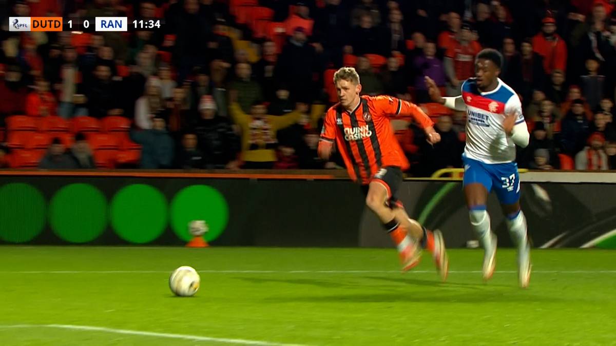 Dwóch piłkarzy walczy o piłkę na zielonym boisku. Zawodnik w pomarańczowej koszulce biegnie w stronę kamery, a za nim podąża zawodnik w białej koszulce.