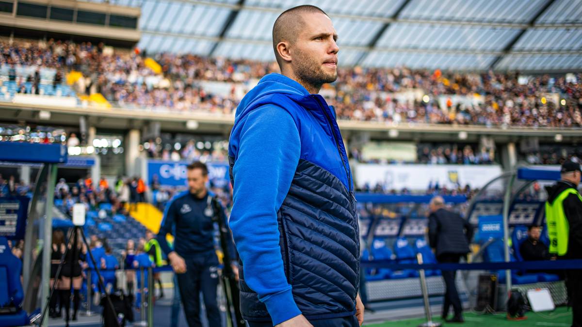 Mężczyzna w niebieskiej kurtce z kapturem i kamizelką stoi na tle trybun stadionu piłkarskiego.