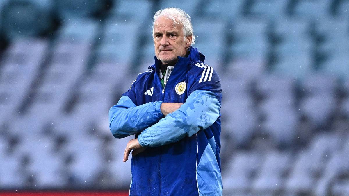 Mężczyzna w średnim wieku, ubrany w niebieską kurtkę sportową z białymi paskami, stoi na tle pustych trybun stadionu.