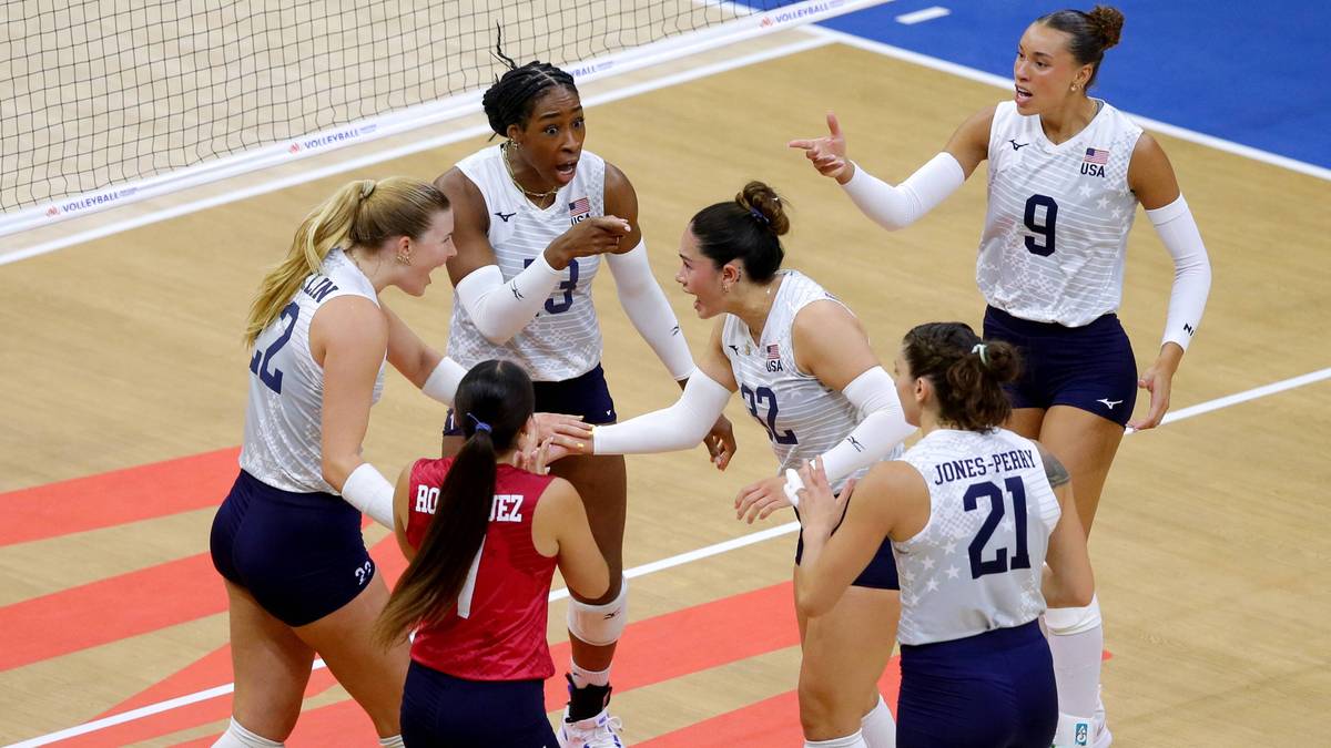 Grupa siatkarek w białych koszulkach i granatowych spodenkach dyskutuje na parkiecie.