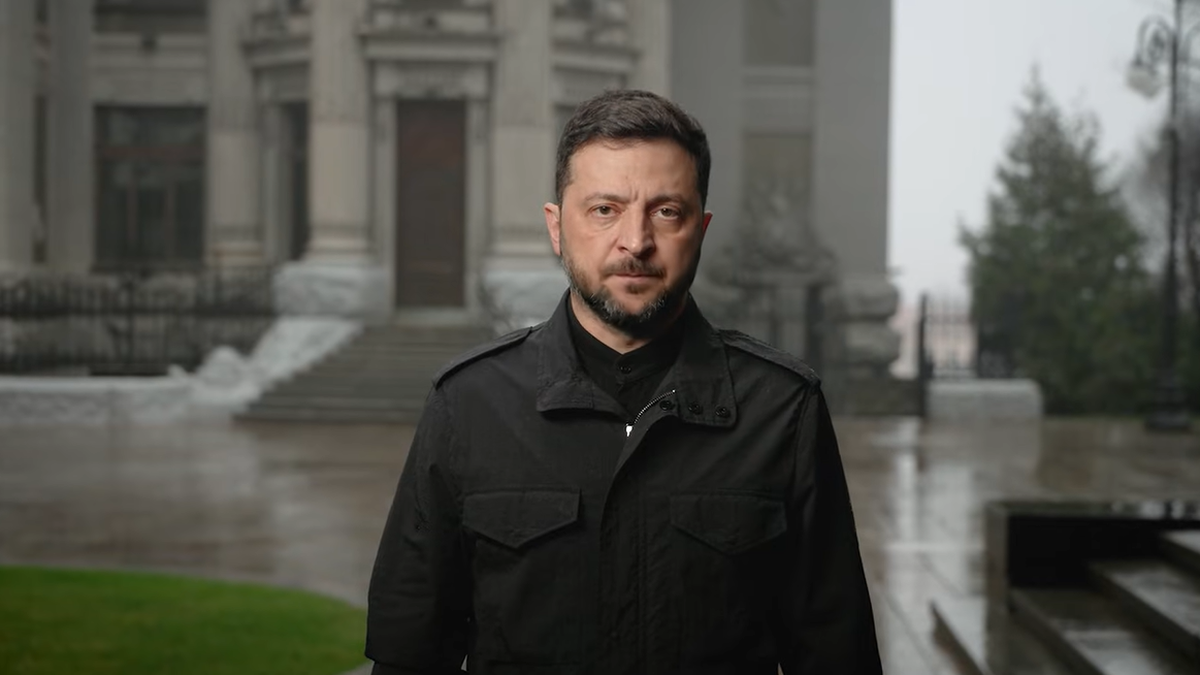 Wołodymyr Zełenski, prezydent Ukrainy, stoi przed budynkiem w deszczowy dzień.