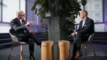 Dw&oacute;ch mężczyzn w eleganckich garniturach siedzi na krzesłach w nowoczesnym wnętrzu i rozmawia. Jeden z mężczyzn gestykuluje dłońmi podczas wypowiedzi.