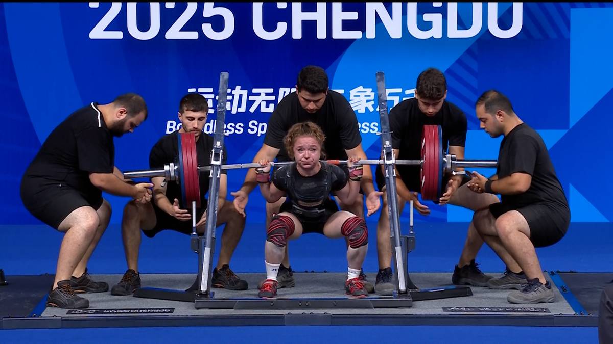 Dziewczynka w pozycji przysiadu z podnoszoną sztangą w centrum, otoczona przez pięciu mężczyzn w strojach sportowych. Z tyłu widoczne logo '2025 CHENGDU'.