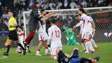 Piłkarze w białych strojach cieszą się po strzelonej bramce na zielonym boisku piłkarskim.