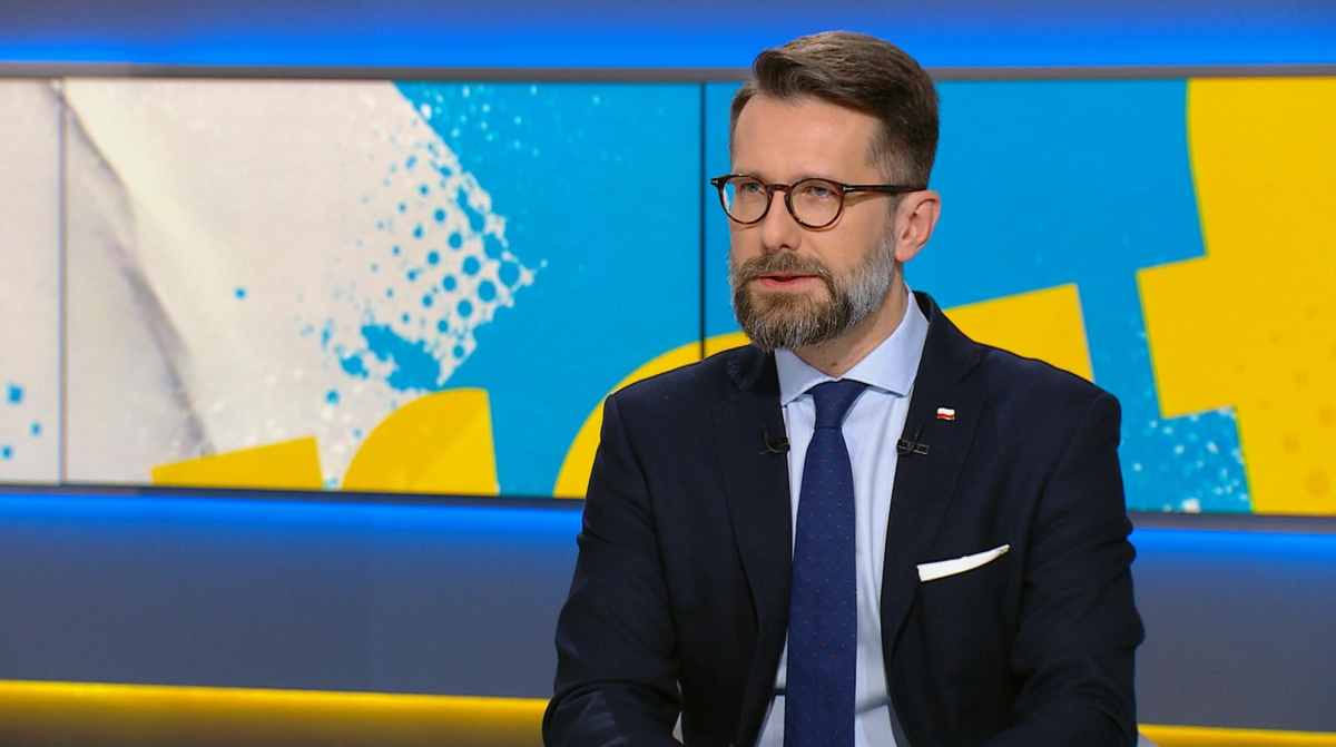 Mężczyzna w okularach i garniturze siedzi w studiu telewizyjnym.