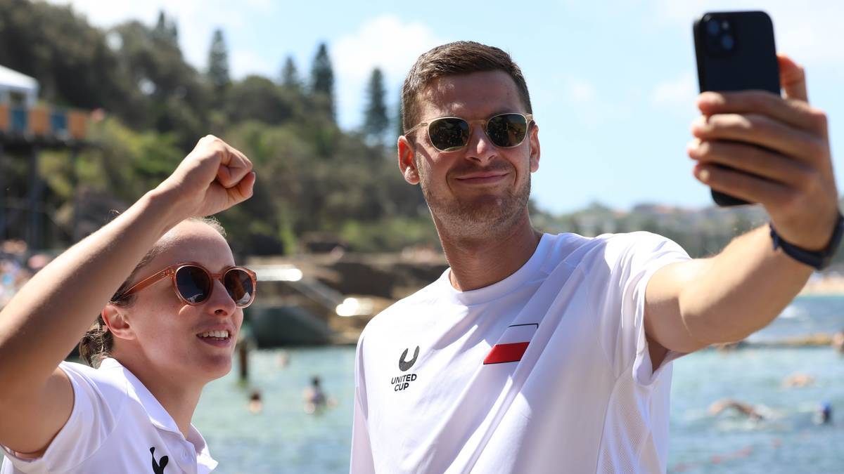 Hubert Hurkacz i Magda Linette pozują do selfie na plaży, oboje uśmiechają się do aparatu. Hubert trzyma telefon w prawej dłoni, a jego lewa pięść jest uniesiona w geście radości.
